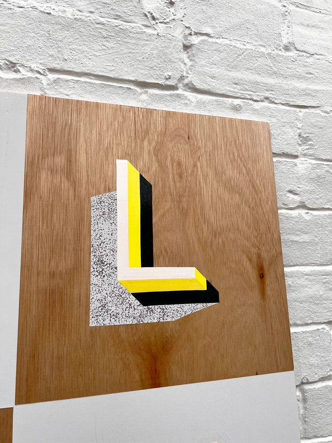 Close up of letter L from PLAY hand painted sign in neon yellow, lilac and grey, against a white brick wall, at an angle. 