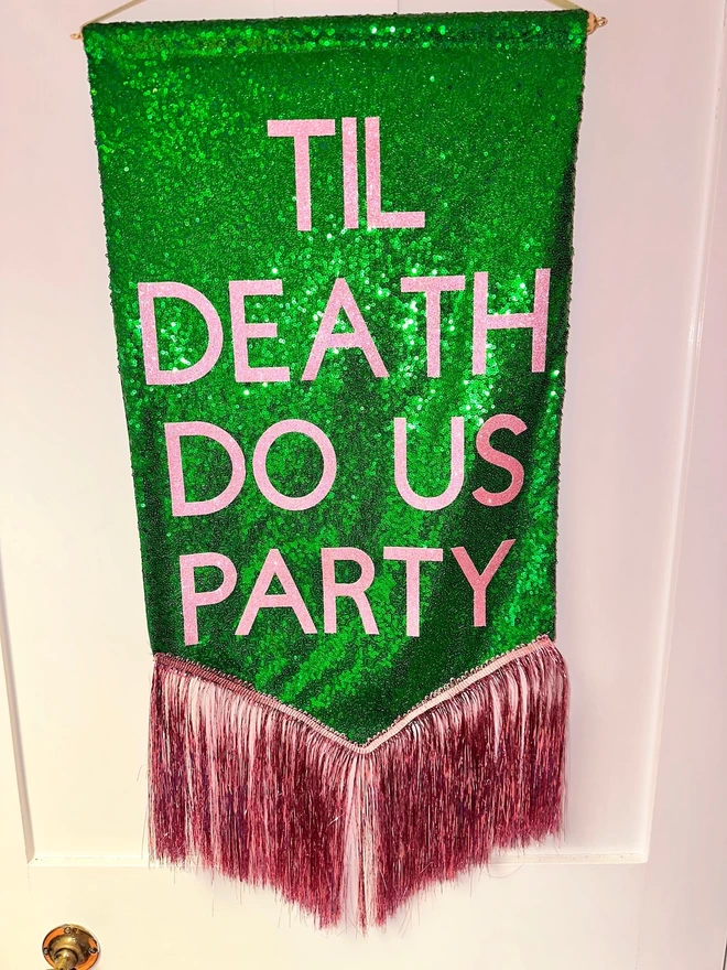  midi customisable banner hangs from a hook. It has a green sequin background and a light pink tinsel trim along the bottom. The text is light pink and says 'TIL DEATH DO US PARTY'
