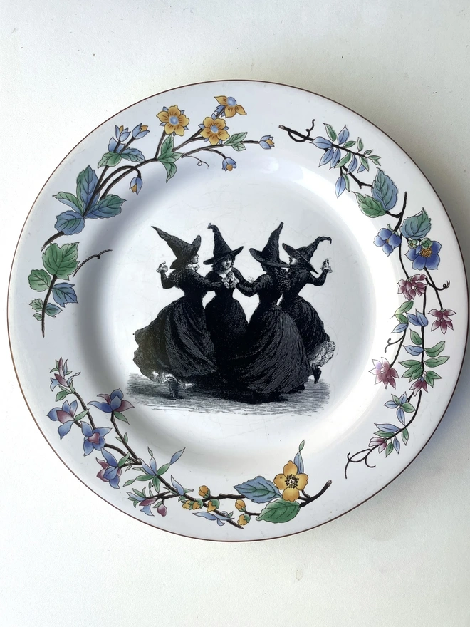 a coven of 4 witches dance hand in hand, the image is victorian in black and white and is printed directly onto an ornate vintage plate