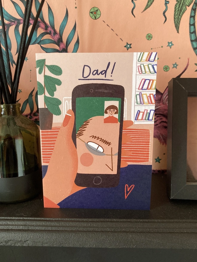 Dad FaceTime card is on a mantlepiece, with pink wallpaper in the background
