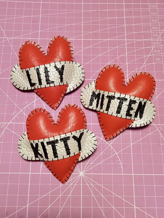 Three red heart brooches placed on a pink cutting mat, showing the names LILY, MITTEN and KITTY in black letters on white scrolls