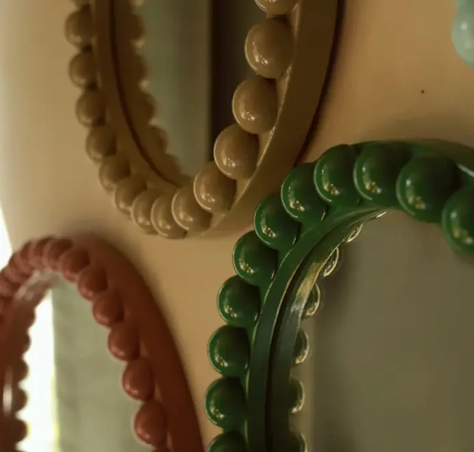Many round oversized bobbin mirrors, in different colours and sizes with a decorative bobbin-style wooden frame, hanging on a white wall.