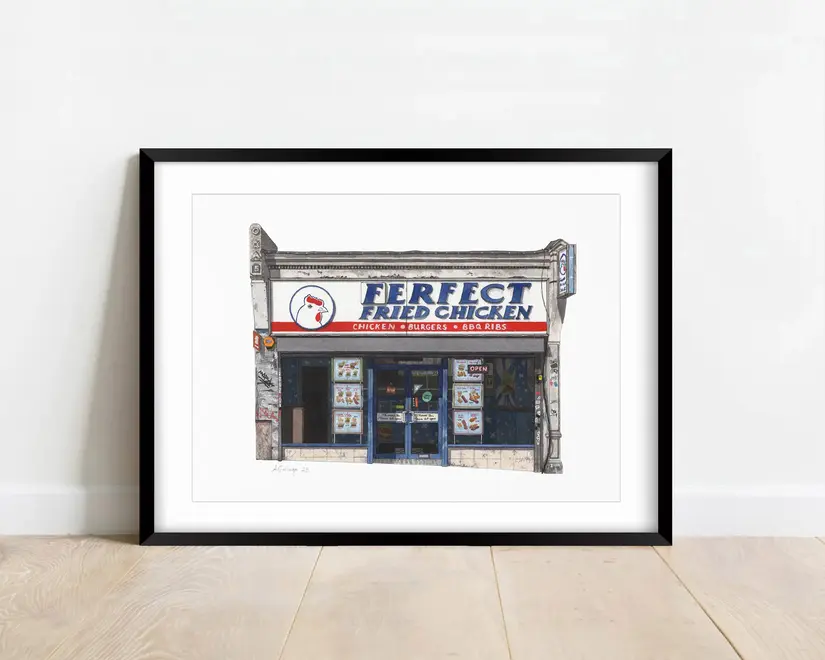 Forest Hill Ferfect Fried Chicken Shop Giclée Print, a print of a fried chicken shop in a black frame on a wooden surface against a white backdrop. 