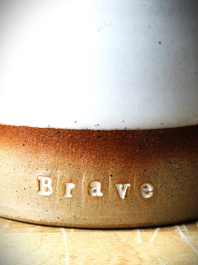 Rustic ceramic mug with a white speckled upper glaze and brown lower half, featuring the word “Brave” on the front, sitting on a wooden table against a neutral background.