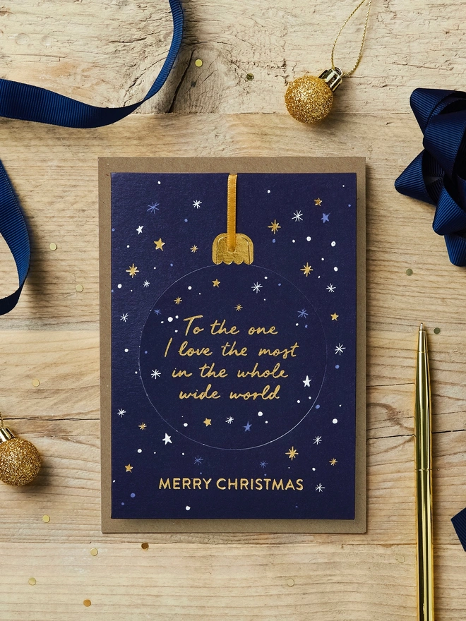 Christmas pop-out bauble card with "To the one I love most in the whole wide world" on it sat on a wooden desk with blue and gold Christmas decorations and stationery.