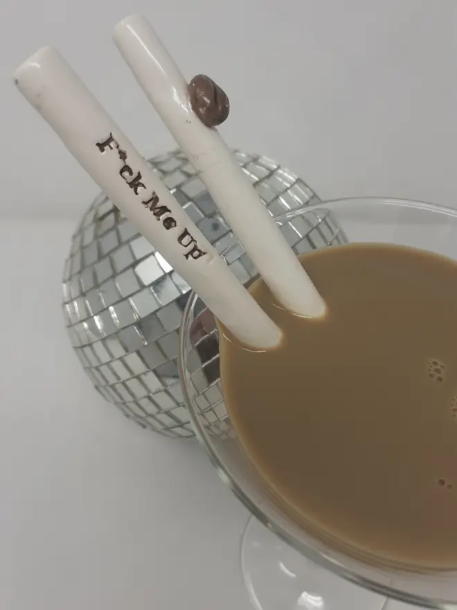 Two ceramic straws in a glass of Espresso Martini, One straw has the words "F*ck Me Up" in coffee brown and the other is decorated with a coffee bean. There is a mirror ball in the background.