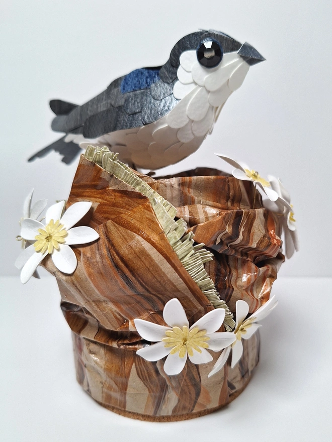 Housemartin bird sculpture, perched on a floral tree stump. This handmade sculpture is made from recycled paper. The bird is black and blue, with a white belly. White and yellow flowers adorn the tree stump. Side view.