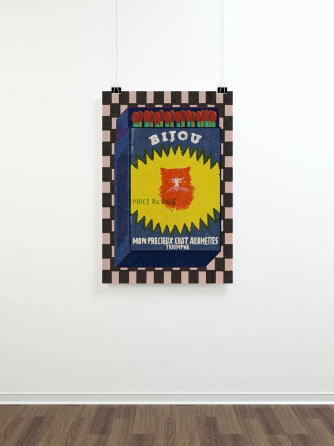 An illustrated vintage-style matchbox labeled “BIJOU.” The box is dark blue with a jagged yellow burst shape in the center, featuring a bright red cat’s face. Text on the burst reads “PRICE Re. 0.06.” Along the bottom, white lettering says “MON PRECIEUX CHAT ALLUMETTES TRIOMPHE.” The top of the matchbox shows red-tipped matches with green stems. The background is a checkered pattern in black and white.