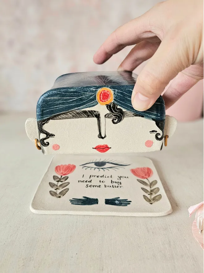 A whimsical ceramic butter dish shaped like the head of a fortune teller, painted in an illustrative style. The fortune teller wears a turban with a few curls escaping, and has a mystical expression. The base of the dish features delicate flowers, an all-seeing eye, and the playful words “I predict you need to buy some butter,” adding a quirky touch of fortune-telling flair to your kitchen.