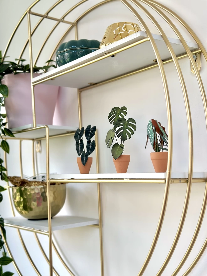 A miniature replica Monstera Deliciosa paper plant ornament in a terracotta pot with another paper plant either side sat on a shelf of a gold circle shelving unit with other gold and pink ornaments
