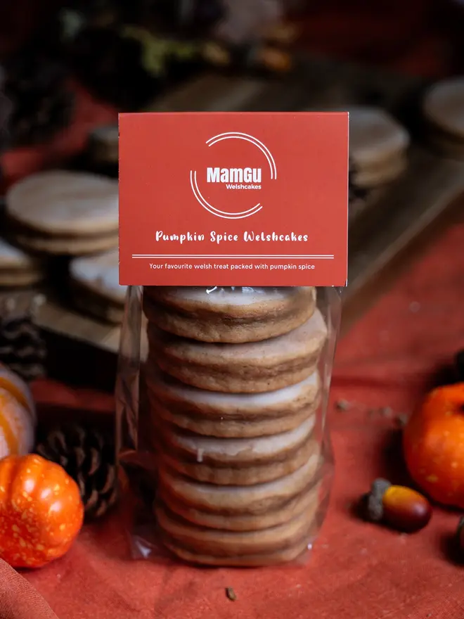 6 pumpkin spice welsh cakes in packaging with an orange label on top. in the background there are Pumpkin Spice Welsh Cakes on a wooden chopping board, an orange table cloth, and surrounded by autumnal decorations such as pumpkins and pine cones.