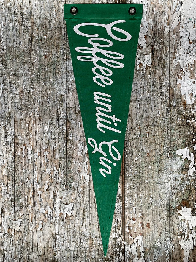A green pennant flag hung vertically on a wall with the words 'Coffee before Gin' written in ivory canvas 