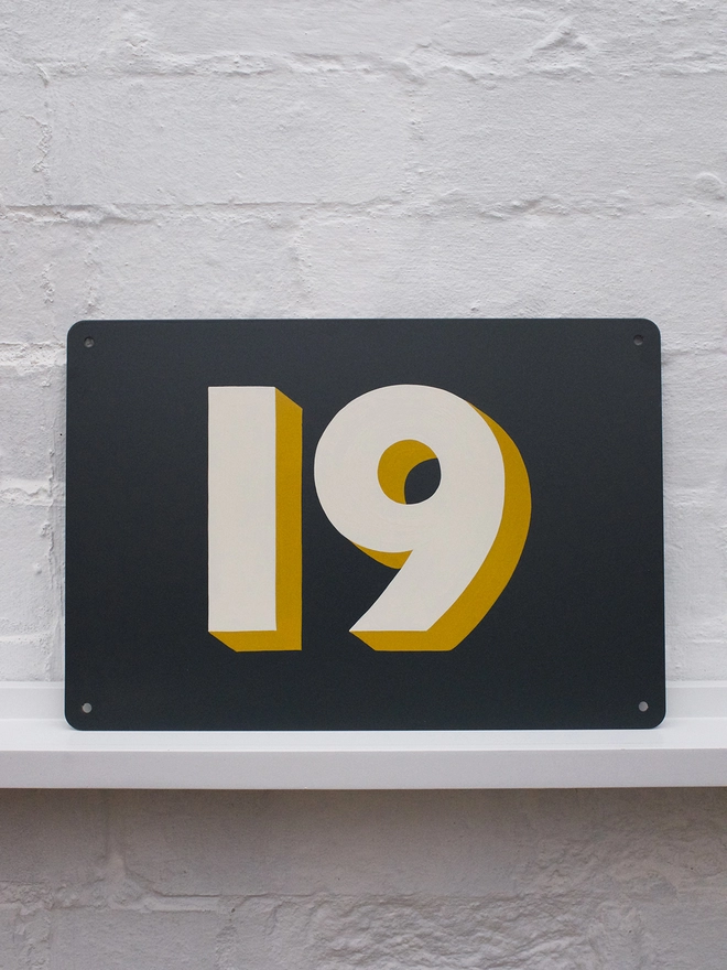 Hand painted off white and mustard house number 19 on an anthracite grey metal plaque against a white brick wall. 