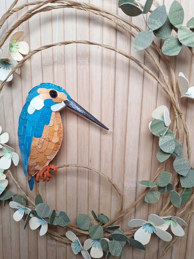 Close up of a handmade kingfisher bird. The paper sculpture is blue, orange and white, with a long grey beak. The kingfisher sits on a wreath of peach tree blossom.