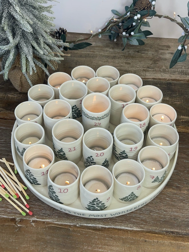 alternative advent tray with 24 numbered candle holders on a rustic table top with christmas tree and matches