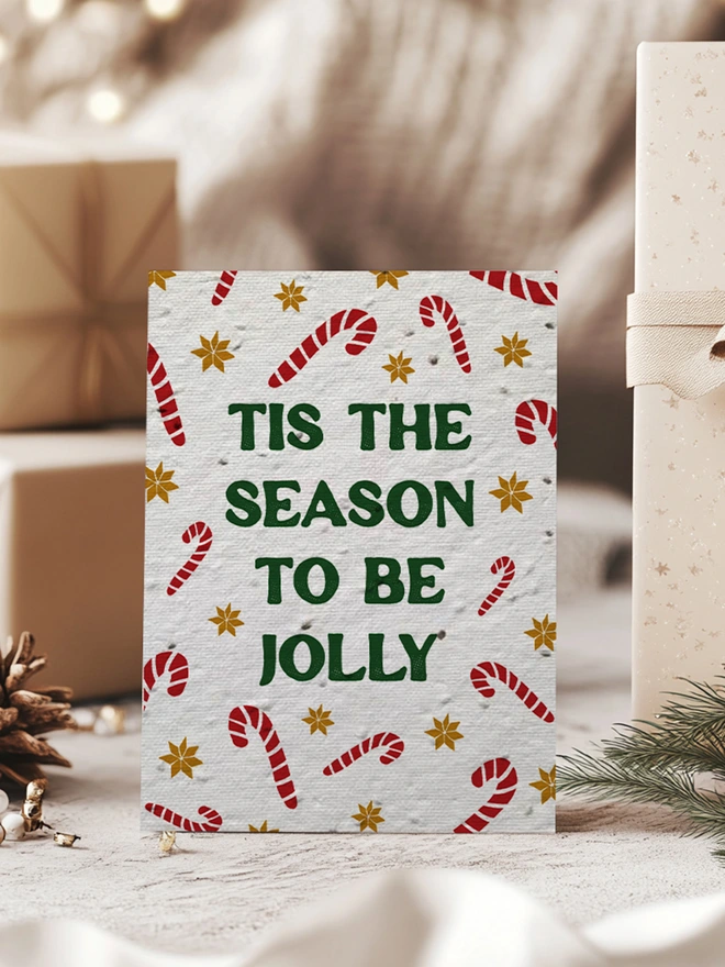 Christmas Card with candy canes and gold star pattern with 'Tis the season to be Jolly' in bold forest green text. The card is surrounded by Kraft paper wrapped presents.