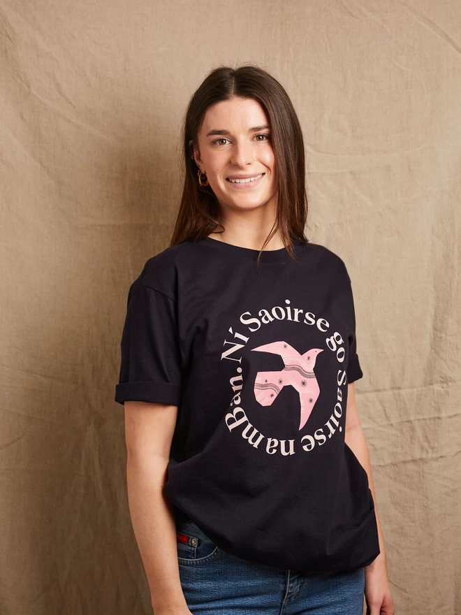 Model is shown wearing a Black & Beech navy heavyweight t-shirt with the words Ní Saoirse Go Saoirse Na mBan in pink written in a circle surrounding a pink bird