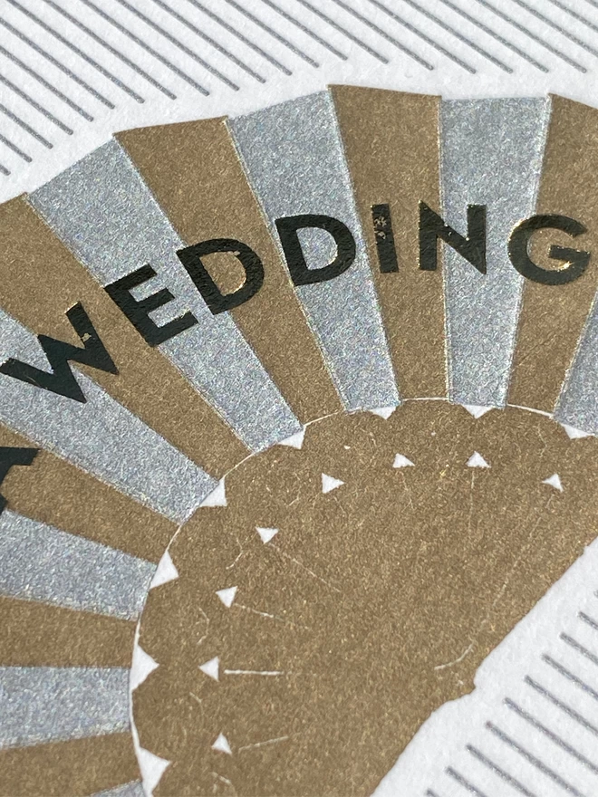 Close up of aWhite and silver striped card with silver and gold fan on it and gold text reading 'Wedding'.