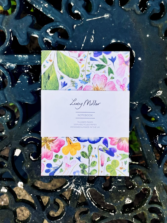 Compact Pocket-Sized Notebook with Pressed Flower Design, 'Lucy Miller' Branded Belly Band, Laying Flat on Wrought Iron Garden Table