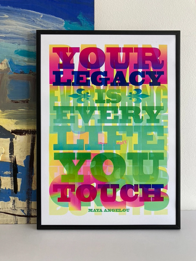 Framed multicoloured typographic print of a Maya Angelou quote: “Your Legacy is every life you touch in rainbow colours”. This is printed over the E.E. Cummings quote “Damn everything but the circus” - also appropriated by Corita Kent.  The print rests against a blue and yellow abstract painting.