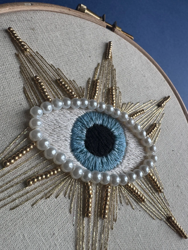 A close up of an embroidery hoop on a blue background containing a hand stitched protective eye edged with pearl beads surrounded by a stitched gold star and gold beads