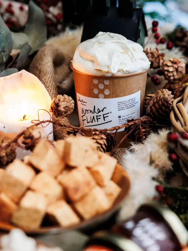 Vanilla Wonder Fluff Tub, marshmallow fluff on a hot chocolate. This is surrounded by lots of tasty treats on a table. 