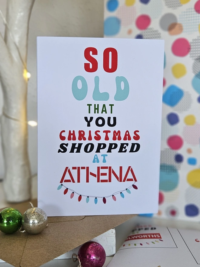 A photograph of the front of a humorous A6 greeting card illustrated using brightly coloured text and the retro shop logo. The text says so old  that you Christmas shopped at Athena with string lights