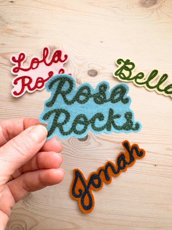 A chain stitched embroidered patch in teal and green spelling Rosa Rocks being held up in front of 3 other personalised patches