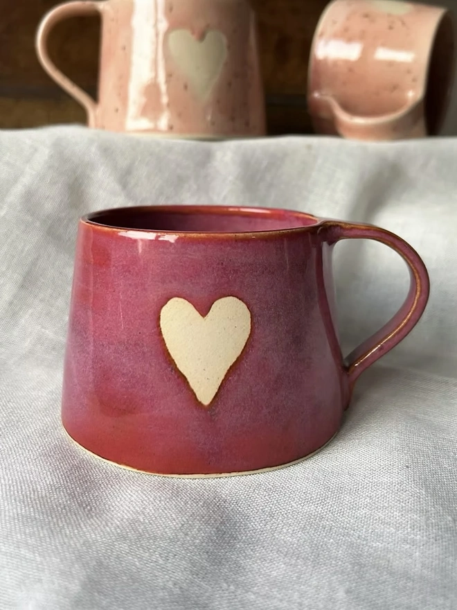 Heart Mug Raspberry Red, red heart mug on a white surface in front of a pair of pink heart mugs. 