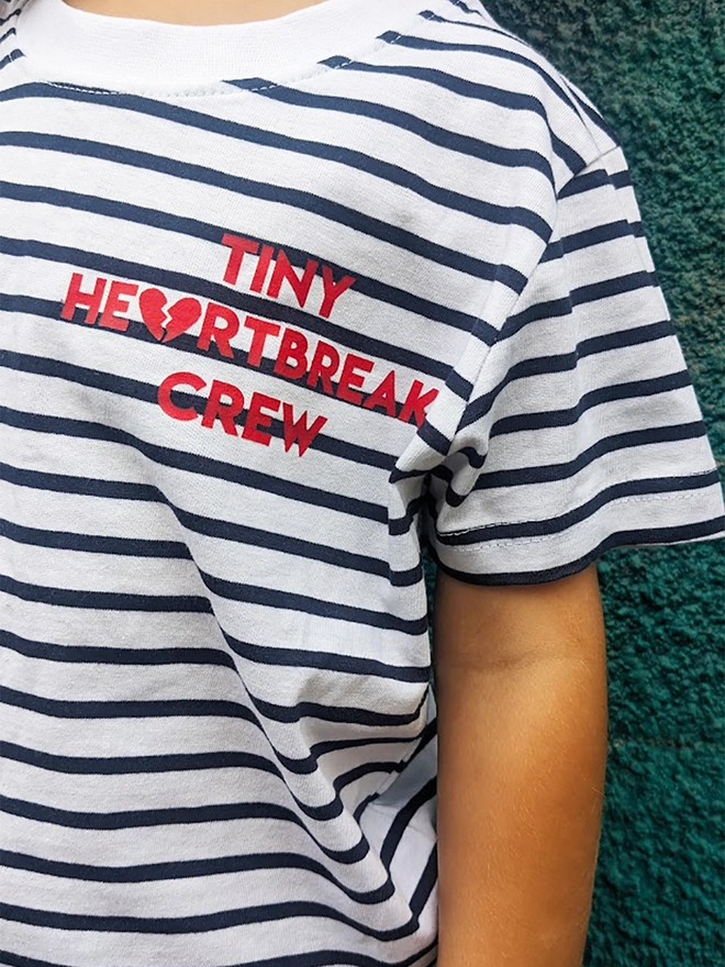 Child wearing a navy and white stripe t-shirt with red Tiny Heartbreak Crew slogan.