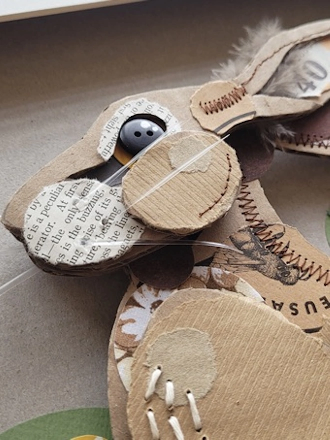 Startled Hare, close up of face showing the varied materials used to create the art piece. Buttons for eyes, plastic straw pieces for whiskers and a variety of torn brown papers to create a fur effect. Hand sewn white chest hairs.
