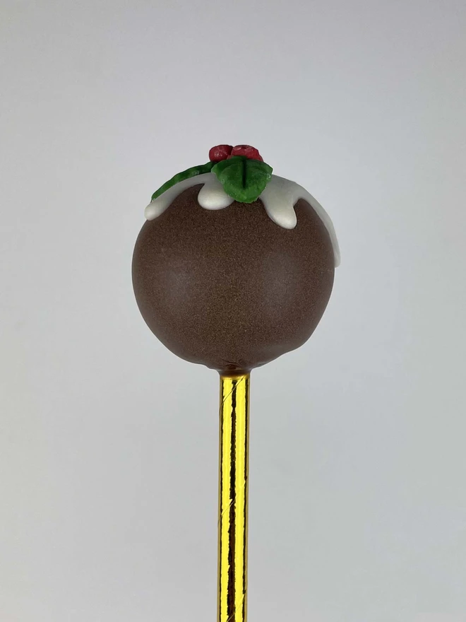 A single Christmas Pudding Cake Pop (Pack of 6, 12, 18, 24)  in a white chocolate drip and topped with holly made from fondant.