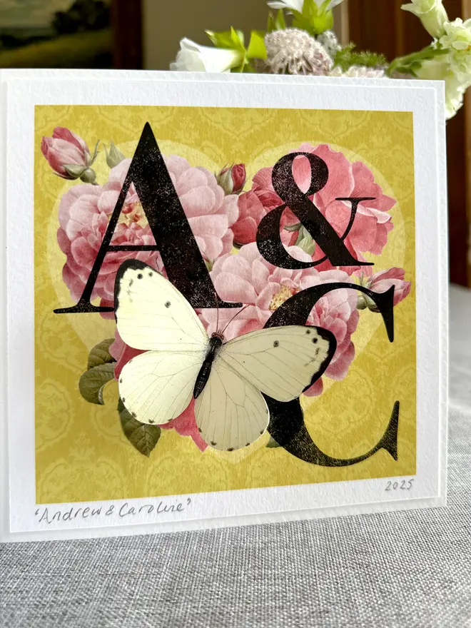 A hand made Golden wedding anniversary card featuring a heart made of deep pink roses on a golden background with letters and initials of the couple. A white paper cut three dimensional butterfly sits on the heart.  The couple's names are hand written and signed on the bottom left hand corner with pencil. The butterflygram card sits in a home setting. 