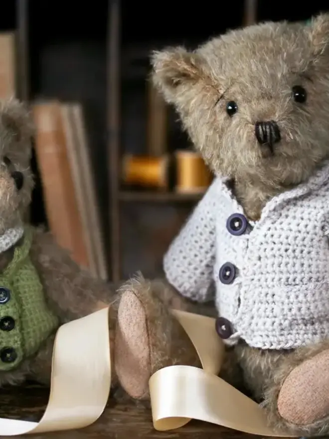Brown teddy bear sitting upright on a bed in room, wearing a cream knitted cardigan with three buttons.