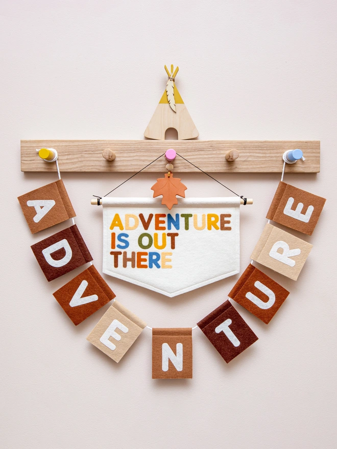 White felt banner with the words "Adventure is out there" written on it in a mix a earthy, jungle themed colours. Surrounding the banner is a felt bunting in a range of brown colours that also says the word "adventure"