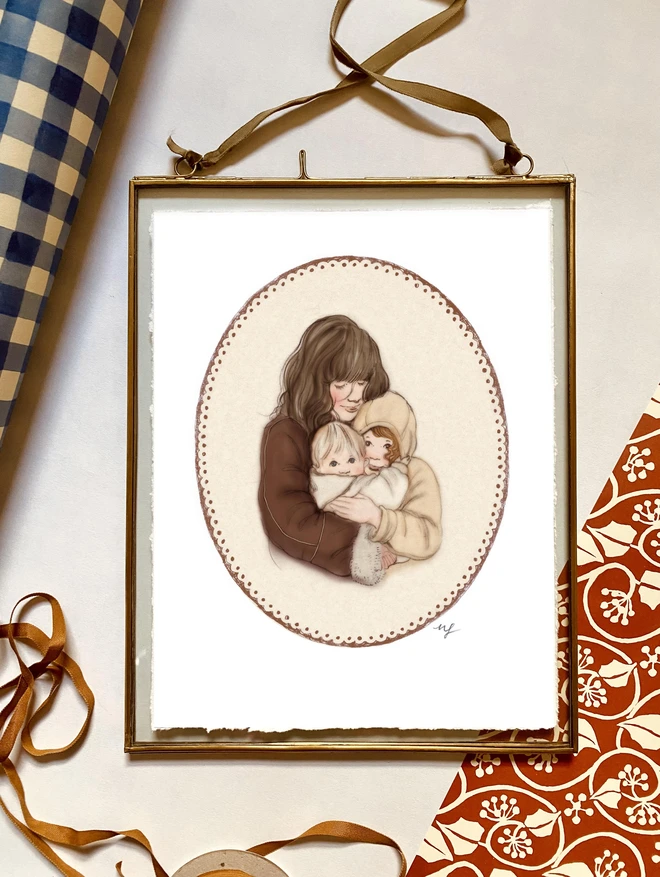 a photograph of a framed personalised print of a mother hugging her 2 children. The print is vintage illustration in style and the frame is glass with a brass edge and velvet ribbon hanging