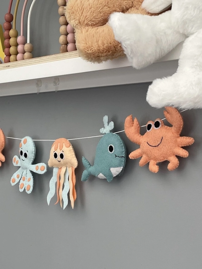 Nursery Under The Sea Garland, a sea themed garland hanging from a white shelf against a grey backdrop in a child's bedroom. 