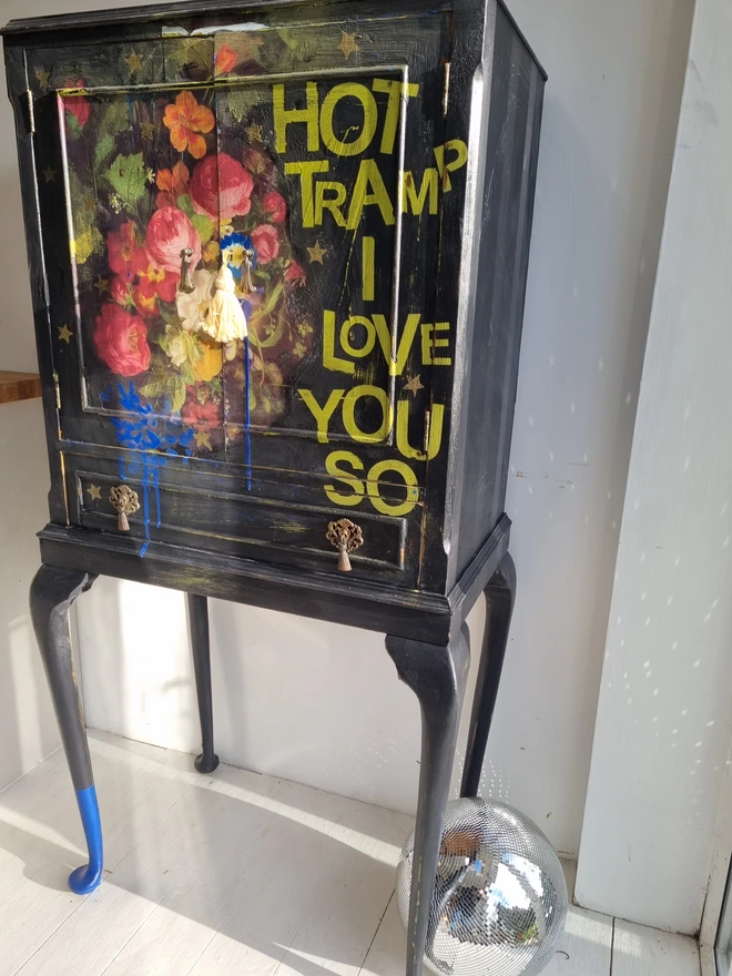 Elegant cocktail cabinet in black with colourful florals and text in chartreuse green that reads 'hot tramp, I love you so', a David Bowie lyric. Heavily distressed to age it. The interior is cobalt blue.