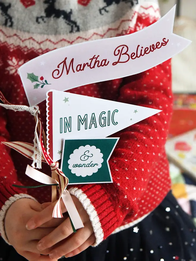 A child wearing a traditional red Christmas jumper holds three personalised Christmas flags made from cardboard. Each flag is attached to a wooden stick with ribbons. The flags read "Martha Believes in magic & wonder".