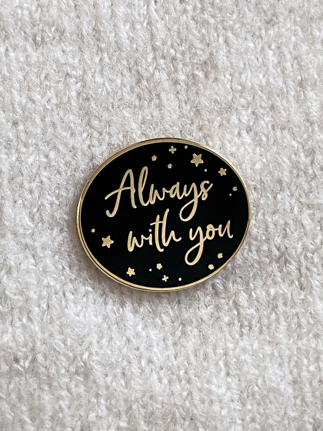 A black and gold enamel pin badge is pinned to a beige jumper. It has a subtle star design and the words "Always with you".