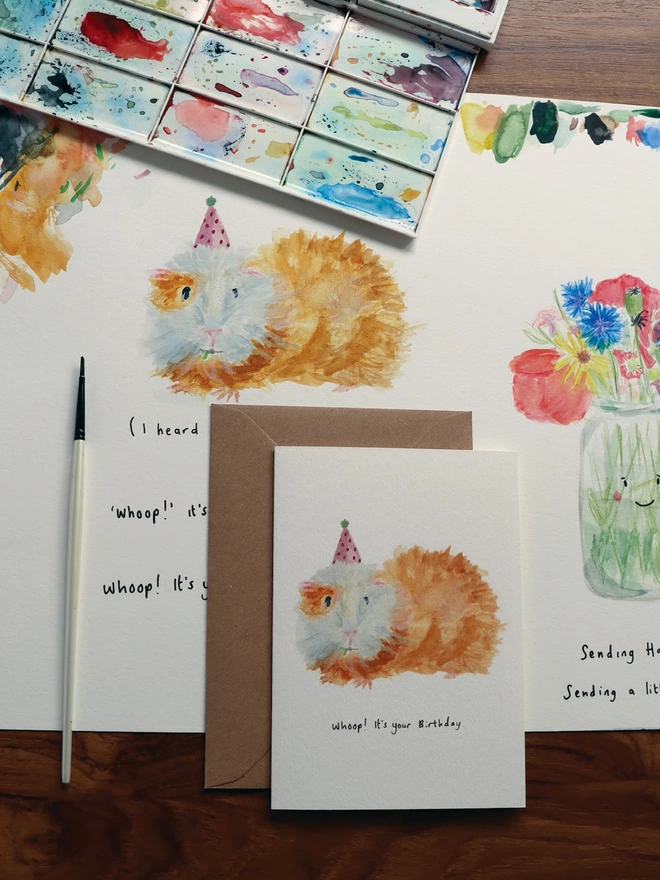 Desk Shot Of The Guinea Pig Greeting Card Sitting Along Side The Original Hand Painted Watercolour Illustration, Paintbrush and Palette