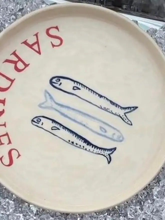 'Sardines' Red Wording Ceramic Side Plate With 3 Fish, a ceramic side plate sitting on ice with a fish design. 