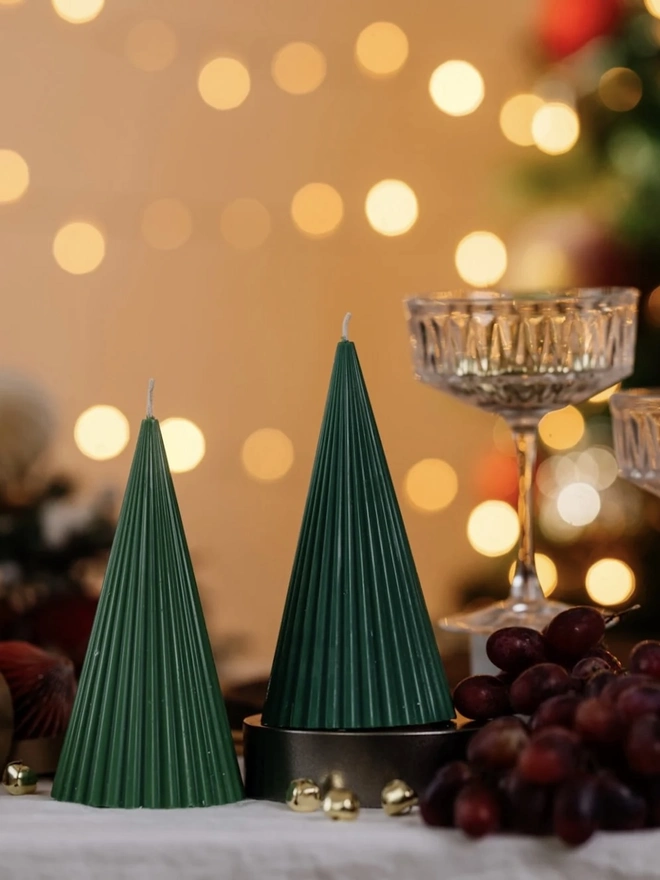Green Christmas Tree Cone Candles. Two forest green ribbed cone candles, one raised on a dark stand, beside crystal coupe glasses and red grapes on a linen tablecloth. The fine vertical ridges give these minimalist tree candles an elegant, festive look.