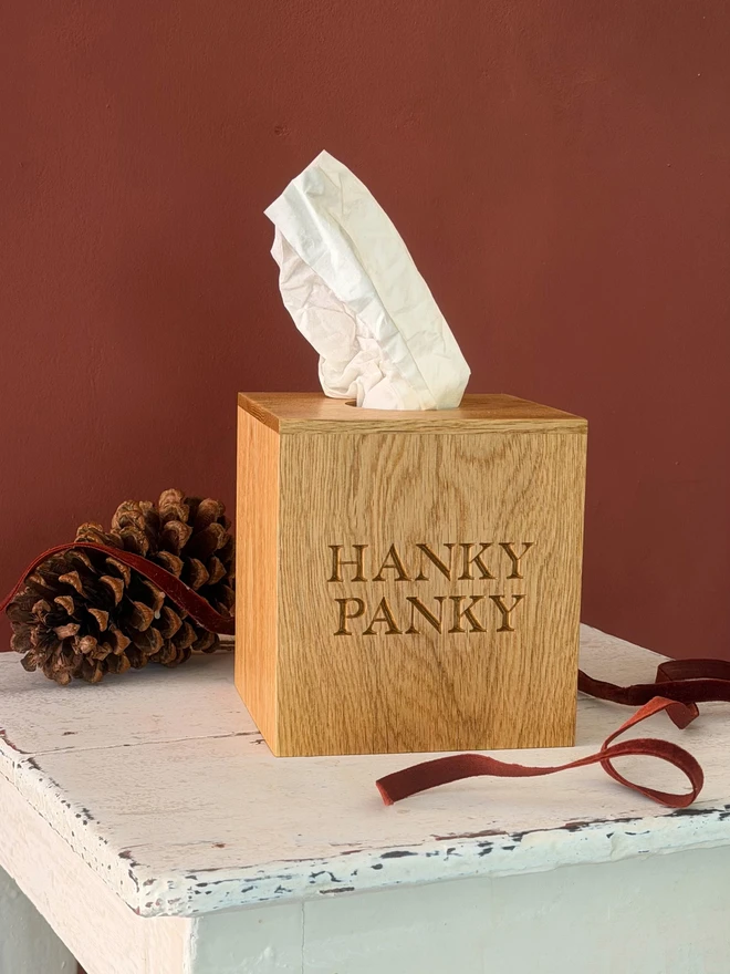 Oak tissue box cover engraved HANKY PANKY, on a white painted stool against a red wall