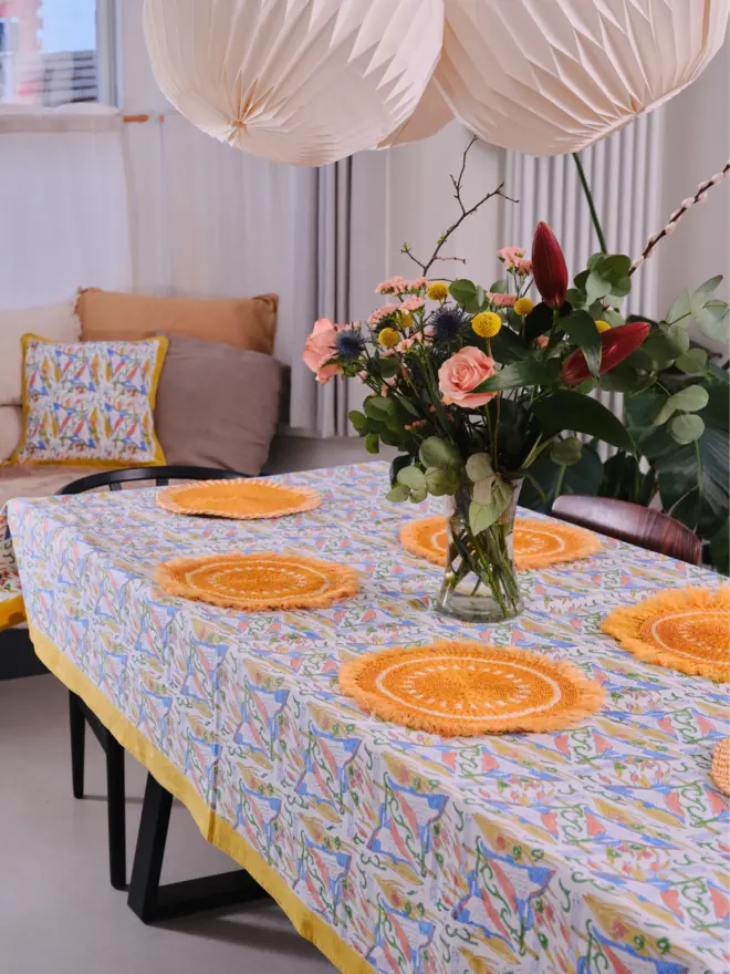 A yellow fringed placemat sitting on top of a colourful tablecloth