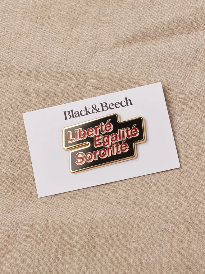 A black enamel pin with a gold edge with the words Liberté Egalité Sororité written in red with a white boarder, on a white backing card 