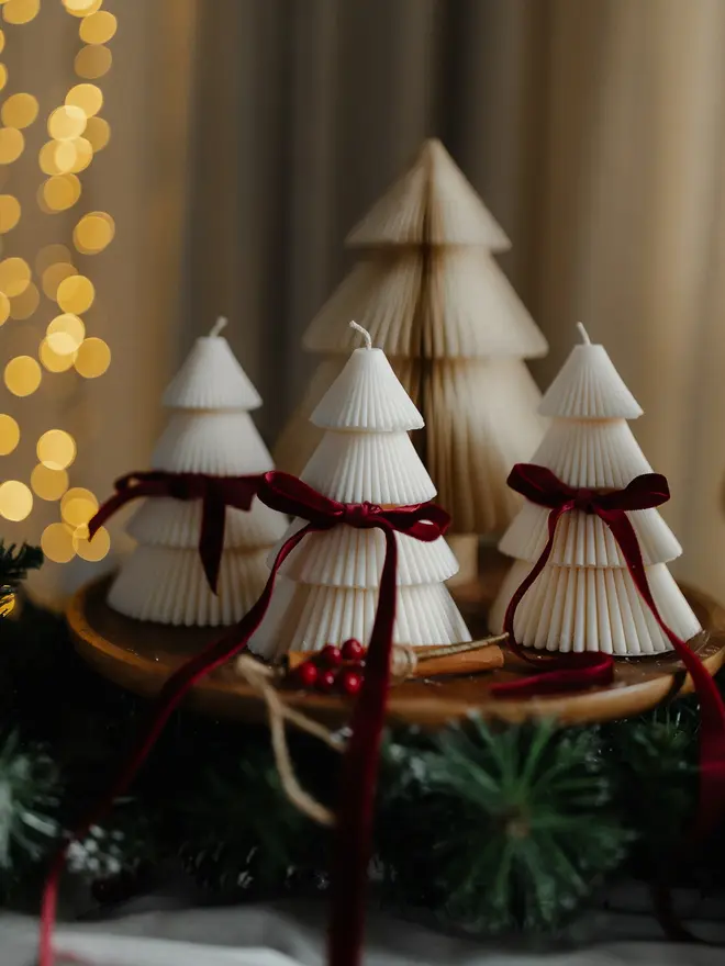 Tiered Christmas Tree Candle. Three lit cream Christmas tree candles with ribbed tiers and burgundy velvet bows, arranged on a wooden board in front of a paper honeycomb tree. Pine garland, cinnamon sticks and red berries complete this cosy festive centrepiece.