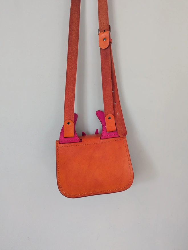 Orange leather monster cross body bag hanging against a blue wall, rear view.