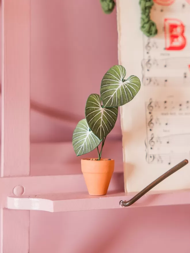 A miniature replica Anthurium Regale paper plant ornament in a terracotta pot balanced on the end of a leaf of a giant Bird of Paradise plant with other plants in the background
