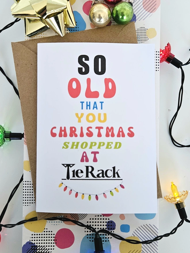 A photograph of the front of a humorous A6 greeting card illustrated using brightly coloured text and the retro shop logo. The text says so old you Christmas shopped at Tie Rack with a string of fairy lights
